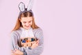 Girl in rabbit bunny ears on head with basket full of colored eggs on pink background. Cheerful smiling happy child. Easter