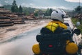 A girl in a raincoat, shoe covers and a helmet is sitting on big adventure motorbike. Rain and felling after the storm. Sawmill.