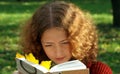 Young curly girl reads a book in a city park. The book contains a bright yellow autumn leaf Royalty Free Stock Photo