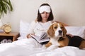 A girl is reading a book in bed under a blanket. A beagle dog is nearby. Royalty Free Stock Photo