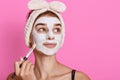 Girl receiving cosmetic white facial mask, looking dreamily aside, wearing hairband with bow, charming lady doing anti aging Royalty Free Stock Photo