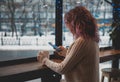 Girl with red curly hair in a white sweater sits in a cafe opposite a large window