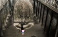 A girl in a red dress is sitting at the feet of the Buddha and praying. There is a very grand and linear statue of Buddha, Ellora