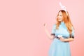 A girl with red hair in a blue dress and easter bunny ears on a pink background. A beautiful woman is pointing at a product or Royalty Free Stock Photo