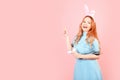 A girl with red hair in a blue dress and easter bunny ears on a pink background. A beautiful woman is pointing at a product or Royalty Free Stock Photo