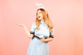 A girl with red hair in a blue dress and easter bunny ears on a pink background. A beautiful woman is pointing at a product or Royalty Free Stock Photo