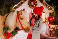 A girl in a red hat strokes a red horse in a Christmas wreath Royalty Free Stock Photo