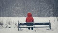 A girl in a red jacket with a hood sits alone, in winter on park benches