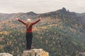 A girl in a red jacket stretching her arms on a mountain, a view of the mountains and an autumn forest by a cloudy day