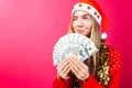 A girl in a red sweater and a Santa hat, holds the money and looks away thinking where to spend it on a red background. Royalty Free Stock Photo