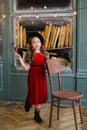 Girl in a red velvet dress holding a red rose, standing near a chair on the the bakery