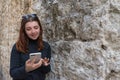 girl relying on stone brick wall of ancient medieval street. Redhead typing smartphone. young woman using cellphone checking the