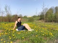 The girl is resting on the spring meadow. She is sitting on the bedspread. Dandelions are blooming, young grass is growing Royalty Free Stock Photo