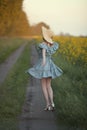 A girl in retro clothes with a straw hat runs along a rural road along a yellow field. Running away into the distance. Royalty Free Stock Photo