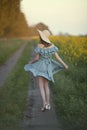 A girl in retro clothes with a straw hat runs along a rural road along a yellow field. Running away into the distance. Royalty Free Stock Photo