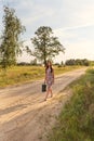 A girl in a retro vintage dress with a suitcase and long flowing hair walks along the road in the yellow rays of the sunlight Royalty Free Stock Photo