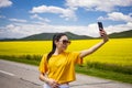 Girl on the road, in a yellow T-shirt, taking a photo in a flowering field, against the backdrop of mountains