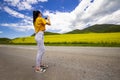 Girl on the road, in a yellow T-shirt, taking a photo in a flowering field, against the backdrop of mountains