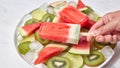 The girl`s hand holds a fruity red popsicle on a stick. A plate with pieces fruit and ice cubes on a gray marble table