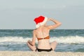 Girl in Santa hat with the inscription New Year on the back is sitting on the beach and looks into the distance