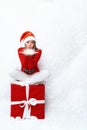 A girl in a Santa hat, sitting on a big red gift box and blowing snow off her palms Royalty Free Stock Photo