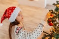 Girl in Santa hat smiling and hanging multi-coloured balls on Christmas tree Royalty Free Stock Photo