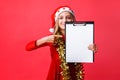 Teen girl in Santa hat and tinsel on neck, holding tablet with clean sheet and pointing finger at him, plans for New year on red Royalty Free Stock Photo