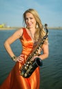 Girl with sax smiles, flirts. Happy blonde woman with big bust plays, performs on goldy saxophone in beautiful dress