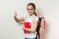 Girl school student, with a backpack and notebooks showing thumbs up sign, background bright wall in school Royalty Free Stock Photo