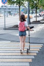 Girl with a scooter in summer in city, crosses road on a zebra crossing, background is granite tiles and asphalt. Short Royalty Free Stock Photo