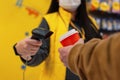 A girl seller in a medical mask from a virus holds a barcode scanner in her hands. A man with a glass of coffee in his hand pays