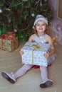 A girl in silver hat sits like a doll near a Christmas tree and holds a gift