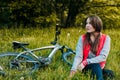 The girl sits in a clearing near the bike. The girl is resting after a walk on the bike