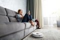 The girl sits on the couch and controls the robot vacuum cleaner using a smartphone Royalty Free Stock Photo