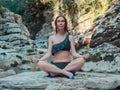 The girl sits cross-legged on a stone by the river, surrounded by rocks. Meditation in nature. Yoga