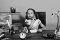 Girl sits at desk with books and clock. Schoolgirl with interested face writes in copybook