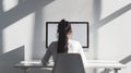 A girl sits at a desktop computer, working against a white, minimalist background with light and shadow, her back