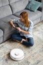 The girl sits on the floor and controls the robot vacuum cleaner using a smartphone, smart home Royalty Free Stock Photo