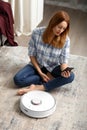 The girl sits on the floor and controls the robot vacuum cleaner using a smartphone, smart home Royalty Free Stock Photo