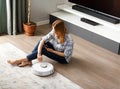 The girl sits on the floor and controls the robot vacuum cleaner using a smartphone, smart home Royalty Free Stock Photo