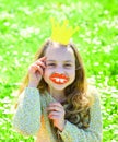 Girl sits on grass at grassplot, green background. Child posing with cardboard smiling lips and crown for photo session