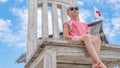 The girl sits on a huge picture chair from below, wearing sunglasses, in summer with blue clouds Royalty Free Stock Photo