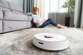 A girl sits in the living room and controls a smart robot vacuum cleaner using a smartphone Royalty Free Stock Photo