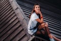 A girl sits on the roof of a house in the city in the evening. Portrait of a model in a dress and sneakers