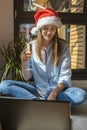 A girl sits in a room on the floor in front of a laptop in a Santa hat and a glass of champagne in her hands and talks Royalty Free Stock Photo