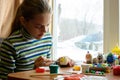 A girl sits at a table by the window and paints Easter eggs with a brush and paints