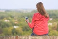 The girl sits on a wall facing her back and using her smartphone