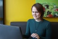 A girl sits, works at a laptop at a table in a cafe on a brown sofa against a yellow wall. On the face is an expression Royalty Free Stock Photo