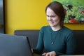 A girl sits, works at a laptop at a table in a cafe on a brown sofa against a yellow wall. On the face is an expression Royalty Free Stock Photo