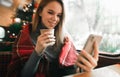 Girl sitting in cozy cafe near window and making photo on smartphone with cup of coffee, cute girl taking selfie on Christmas tree